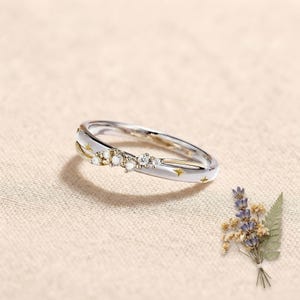 May include: A silver-toned ring featuring a delicate band with a crossover design. The ring is accented with a cluster of small, clear round stones and engraved gold-coloured star motifs along the polished metal surface of the band.