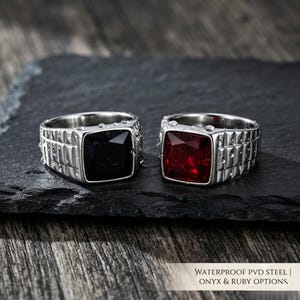 May include: Two silver-toned metal rings with a textured watch-link band design. One ring features a square black stone, and the other features a square red stone. The text WATERPROOF PVD STEEL | ONYX & RUBY OPTIONS is visible in the corner.