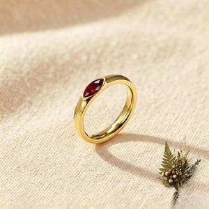 May include: A gold band ring featuring a single marquise-cut deep red gemstone set horizontally into the metal. The ring has a smooth, polished finish and a rounded profile, showcasing a minimalist design with a vibrant, faceted center stone.