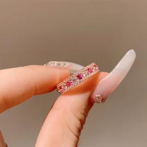 Puede incluir: Un anillo de banda en oro rosa con una fila central de piedras redondas alternas en rosa y transparente, bordeada por dos filas delgadas de diminutos cristales transparentes en pavé. El metal tiene un acabado pulido y diseño simétrico.