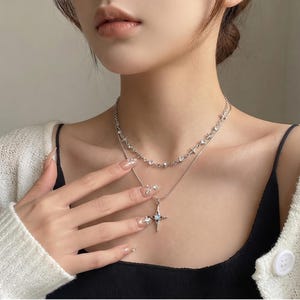 Puede incluir: Un collar de capas en tono plateado con dos cadenas. La cadena más corta está adornada con pequeños dijes en forma de estrella y cristales transparentes. La cadena más larga tiene un colgante central de estrella de cuatro puntas con una piedra azul.