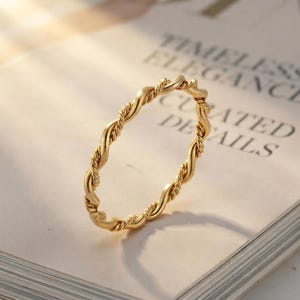 May include: A thin gold ring featuring a continuous twisted rope design. The band has a polished metallic finish and a uniform circular shape, showcasing a detailed spiral texture that wraps around the entire circumference of the jewelry piece.