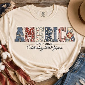 Puede incluir: Camiseta color crema con la palabra "AMERICA" en letras patrióticas rojas, blancas y azules. La camiseta conmemora 250 años con las fechas 1776 y 2026. Un sombrero beige y unos vaqueros azules son visibles en el fondo.