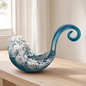 Puede incluir: Una escultura decorativa de vidrio con forma de ola rompiendo. La pieza presenta una base azul translúcida que se transforma en una cresta de espuma blanca opaca y texturizada, con un rizo de vidrio azul liso y alargado hacia la derecha.