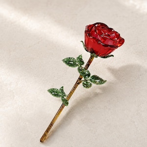 Puede incluir: Una escultura decorativa de rosa de cristal que presenta una flor roja vibrante con pétalos translúcidos, un tallo transparente y hojas de vidrio verde detalladas unidas al tallo. La pieza está elaborada con superficies lisas, reflectantes y texturas.