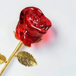 以下が含まれることがあります： 金色の茎と葉が付いた赤いガラスのバラ。バラは鮮やかな赤色で、複雑なディテールがあります。茎と葉は金色で、エレガントさを添えています。バラは装飾品です。