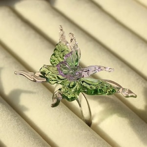 Peut inclure: Une bague ornée d'un papillon en verre translucide. Les ailes du papillon sont principalement vert clair avec de délicates touches violettes près du centre. L'anneau de la bague est transparent et lisse, soutenant la figurine en verre.