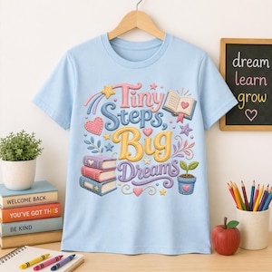 May include: Light blue t-shirt with the phrase "Tiny Steps, Big Dreams" in colorful, embroidered-style lettering. The design includes books, hearts, stars, and a potted plant. The shirt is hanging on a wooden hanger.