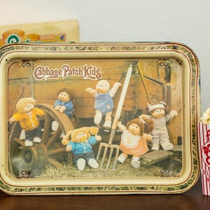 May include: A vintage Cabbage Patch Kids metal tray featuring the dolls. A box with the Cabbage Patch Kids logo is to the left. A red and white striped popcorn container is to the right.