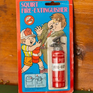 Op de afbeelding: Vintage speelgoed brandblusser op een rode en blauwe kaart. De kaart bevat illustraties van een brandweerman en een rokende man. Het speelgoed is rood met een zwart mondstuk en de tekst "FIRE-EXT".