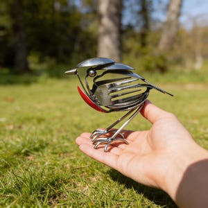 Könnte beinhalten: Eine handgefertigte Metallvogel-Skulptur aus recyceltem Besteck. Der Vogel hat eine rote Brust und einen silbernen Körper. Er wird in einer Hand gehalten, mit einem Hintergrund aus grünem Gras und Bäumen. Ein kreatives Kunstwerk.