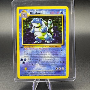 May include: A Blastoise Pokémon trading card, stage 2, with 100 HP. The card features a blue and white turtle-like creature with the text "Blastoise". The card is in a protective case and displayed on a clear stand.