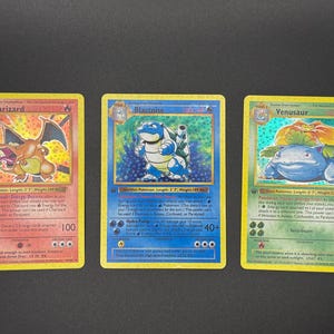 Peut inclure: Trois cartes à collectionner Pokémon : Dracaufeu, Tortank et Florizarre. La carte Dracaufeu présente un dragon orange sur fond de feu. La carte Tortank montre une tortue bleue. La carte Florizarre affiche une créature verte et bleue.