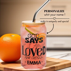 Puede incluir: Un vaso transparente con tapa de bambú y pajita de metal, lleno de una bebida rosa y rodajas de naranja. El vaso presenta texto negro que dice GOD SAYS I AM LOVED AND CHOSEN EMMA, con pequeños iconos de corazones rodeando las letras.