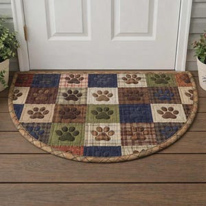 May include: A half-circle doormat with a patchwork design featuring paw prints. The squares are in shades of brown, green, blue, and red plaid. The border is a light brown with a quilted pattern. Placed in front of a white door.