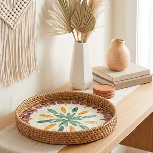 May include: A round woven rattan tray featuring a central mosaic inlay with a floral pattern in shades of green, yellow, and purple. The tray has a high, textured rim and a flat base, showcasing a natural light brown finish on the woven exterior.