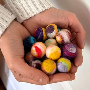 May include: A collection of colorful felt balls in various sizes and hues, including yellow, red, blue, and purple. The balls are held in cupped hands, showcasing their textured, handmade appearance. The background is a soft, neutral tone, highlighting the vibrant colors of the felt balls.