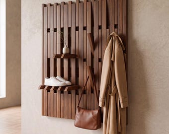 May include: A wall-mounted wooden coat rack featuring vertical slats, integrated shelves, and retractable hooks. A beige trench coat and a brown leather handbag hang from the hooks, while white sneakers and a small vase sit on the shelves.
