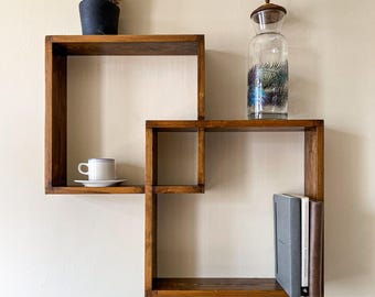 May include: Two interlocking dark brown wooden square shelves mounted on a wall. The top shelf holds a green cactus in a black pot. The bottom shelf contains a white cup and saucer, a glass carafe with a palm leaf print, and three books.