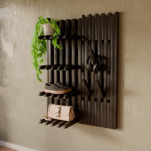 May include: A black wall-mounted wooden slat organizer featuring horizontal shelves and vertical hooks. The unit holds a potted green plant, a pair of gray sneakers, a beige handbag, and black over-ear headphones. The structure is composed of evenly spaced vertical wooden bars.