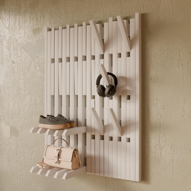 May include: A white wall-mounted organizer featuring vertical slats, integrated hooks, and two small shelves. The unit holds a pair of grey sneakers on the top shelf, a beige handbag on the bottom shelf, and black headphones hanging from a hook.