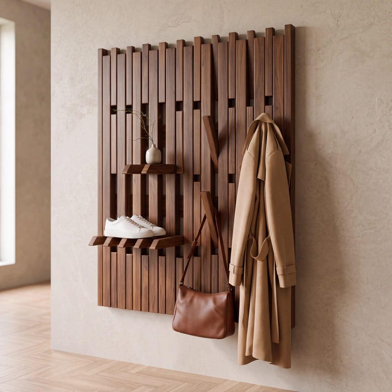 May include: A wall-mounted wooden coat rack featuring vertical slats, integrated shelves, and retractable hooks. A beige trench coat and a brown leather handbag hang from the hooks, while white sneakers and a small vase sit on the shelves.