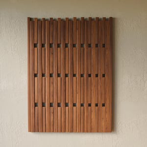 May include: A rectangular wall panel constructed from vertical brown wooden slats of varying lengths, arranged in a repeating pattern with small square gaps between the segments, creating a textured, slatted surface design.