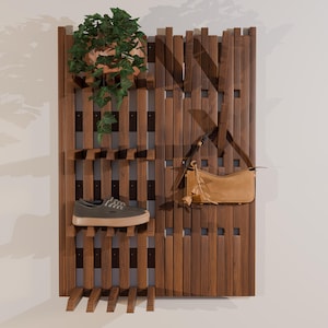 May include: A vertical wooden wall organizer with slats and integrated shelves. A green potted plant sits on the top shelf, a gray sneaker with a tan sole rests on a middle shelf, and a tan leather shoulder bag hangs from a wooden peg.