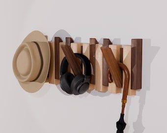 May include: A wall-mounted coat rack featuring alternating light and dark wooden slats. The rack includes several angled pegs holding a tan fedora hat, black over-ear headphones, and a black umbrella with a curved wooden handle.