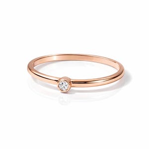 Puede incluir: Un delicado anillo de oro rosa con un solo diamante pequeño y redondo en el centro. La banda es delgada y pulida, reflejando la luz. El anillo se muestra sobre un fondo blanco, resaltando su elegancia simple.