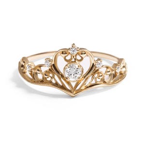 Könnte beinhalten: Ein goldfarbener Ring in Kronenform mit einem zentralen Diamanten und kleineren Diamanten. Der Ring hat ein verziertes, filigranes Design mit einem herzförmigen Mittelteil. Der Ring ist vor einem weißen Hintergrund platziert.