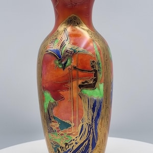 May include: A tall, ornate vase with a reddish-orange background and gold accents. The vase features a detailed painted scene with figures, a bird, and landscape elements in shades of green, blue, and brown. The rim and base are also trimmed in gold.