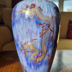 May include: A blue ceramic vase with a gold rim and base, featuring a detailed design of gold dragons with red accents. The vase has a gradient effect, transitioning from a darker blue to a lighter shade.
