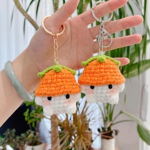 Puede incluir: Dos llaveros de crochet con forma de seta. Cada llavero presenta un cuerpo blanco, una tapa naranja y un ribete verde. Las setas tienen ojos negros y mejillas rosas. Un llavero tiene un anillo dorado, el otro un anillo plateado.