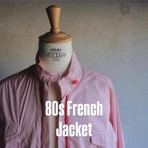 Peut inclure: Veste française des années 80 à rayures roses et blanches, avec une fermeture éclair rouge et des boutons rouges. La veste est présentée sur un mannequin. Le texte "80s French Jacket" est superposé.