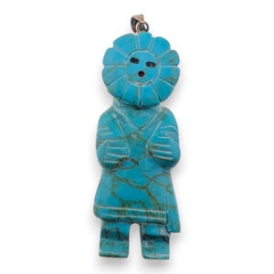 May include: A turquoise pendant carved in the shape of a stylized figure. The figure has a sun-like head with a face, arms crossed at the chest, and a skirt-like bottom. The pendant has a silver-colored bail.