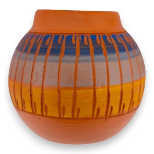 Puede incluir: Un jarrón redondo de terracota con una abertura ancha y acabado mate. El exterior está decorado con bandas horizontales de pintura azul, gris y amarilla, superpuestas con líneas verticales incisas que crean un patrón texturizado y repetitivo.