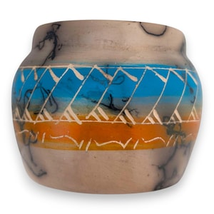 May include: A rounded ceramic pot featuring a matte beige base with black smoke-like markings. A horizontal band wraps around the center, displaying a bright blue and orange gradient overlaid with white etched geometric triangle patterns.