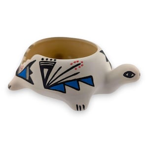 May include: A ceramic turtle-shaped planter with a cream-colored exterior and a brown interior. The turtle's shell is decorated with black, blue, and red geometric patterns. The turtle has a small head and four stubby legs.