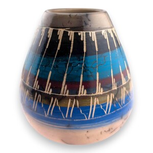 Puede incluir: Un jarrón de cerámica artesanal con forma de lágrima. El jarrón presenta un diseño pintado colorido con bandas horizontales azules, negras y rojas, acentuadas con líneas verticales blancas. La base es de color rosa claro y el borde es gris oscuro.