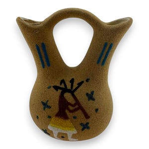 May include: Tan-colored pottery vase with a unique double-spout design and a central handle. The vase features a hand-painted Kokopelli figure in brown, yellow, and white, with blue accents and star-shaped details. A decorative piece.