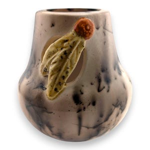 May include: A ceramic vase with a unique design. The vase is primarily a light tan color with dark gray and black streaks. A tan and yellow leaf-like embellishment with a red accent is attached to the side of the vase.