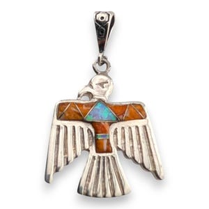 May include: A silver pendant in the shape of a stylized bird with outstretched wings. The bird's body features inlaid orange and blue-green stones. The pendant has a loop at the top for a chain.