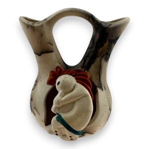 May include: A ceramic vase with a unique design. The vase has a handle and a central opening. It features a figure with red hair and a blue sash. The vase has a speckled, neutral color with dark accents.