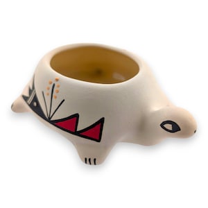 May include: A ceramic turtle-shaped planter in cream with a wide opening. The turtle has black and red geometric designs on its sides and a small eye. The interior is a warm yellow color.