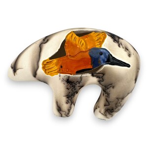 Pode incluir: Uma estatueta de urso em cerâmica com uma superfície lisa em tom esbranquiçado, apresentando padrões de marmoreado escuros e ténues semelhantes a fumo. Um beija-flor laranja e azul brilhante está pintado no lado do urso, em pleno voo.