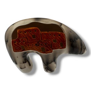 May include: A ceramic bear-shaped sculpture with a hollowed-out interior. The interior features a detailed depiction of a pueblo village in shades of brown. The exterior is a marbled grey and white, with a smooth finish. The sculpture has a unique, artistic design.