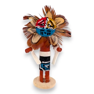 May include: A handcrafted kachina doll figure featuring a round face painted with red, yellow, and blue sections. The head is adorned with brown and tan feathers. The body is cylindrical with a black skirt, white accents, and two white arms.
