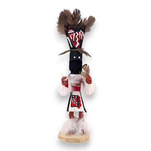 Puede incluir: Una muñeca Kachina nativa americana con cabeza y cuerpo negros, detalles en rojo y blanco y un tocado de plumas. La muñeca sostiene un objeto pequeño y se apoya sobre una base de madera. El diseño de la muñeca incluye patrones geométricos.