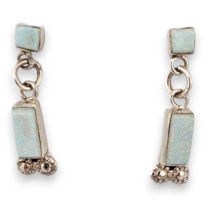 May include: A pair of silver-toned drop earrings. Each earring features a small square top, connected by a chain to a rectangular, light blue stone. The bottom of each earring has a decorative, rounded design.